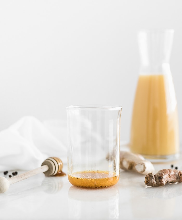 water, honey, grated ginger, grated turmeric, and pepper in the bottom of a glass with orange juice, ginger root, and turmeric in the background.