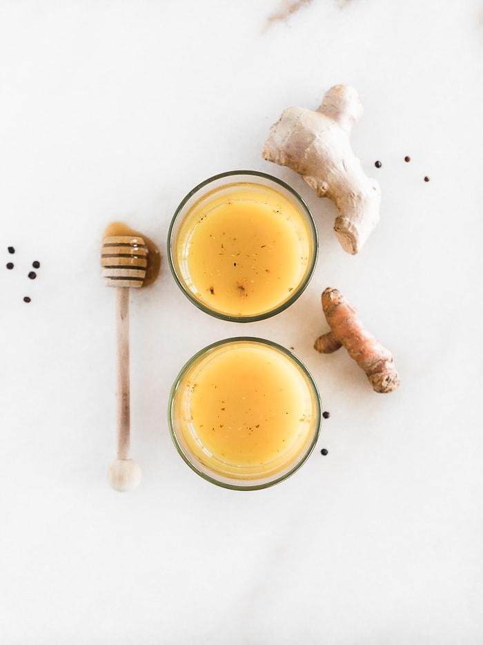 overhead view of two vitamin C shots with a honey dipper, ginger root and turmeric root next to them.