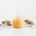 orange juice shot in a glass surrounded by a honey dipper, turmeric root and ginger root.