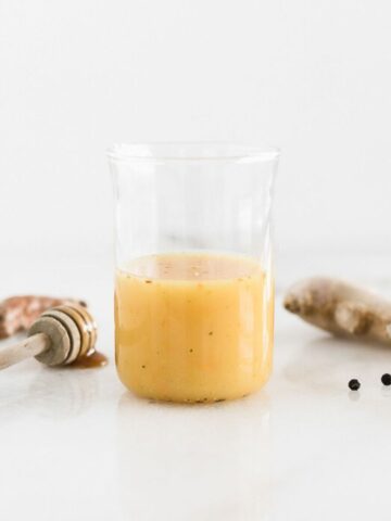 orange juice shot in a glass surrounded by a honey dipper, turmeric root and ginger root.