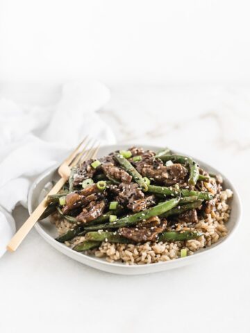 sesame ginger beef and green beans on top of brown rice on a white plate with a gold fork.