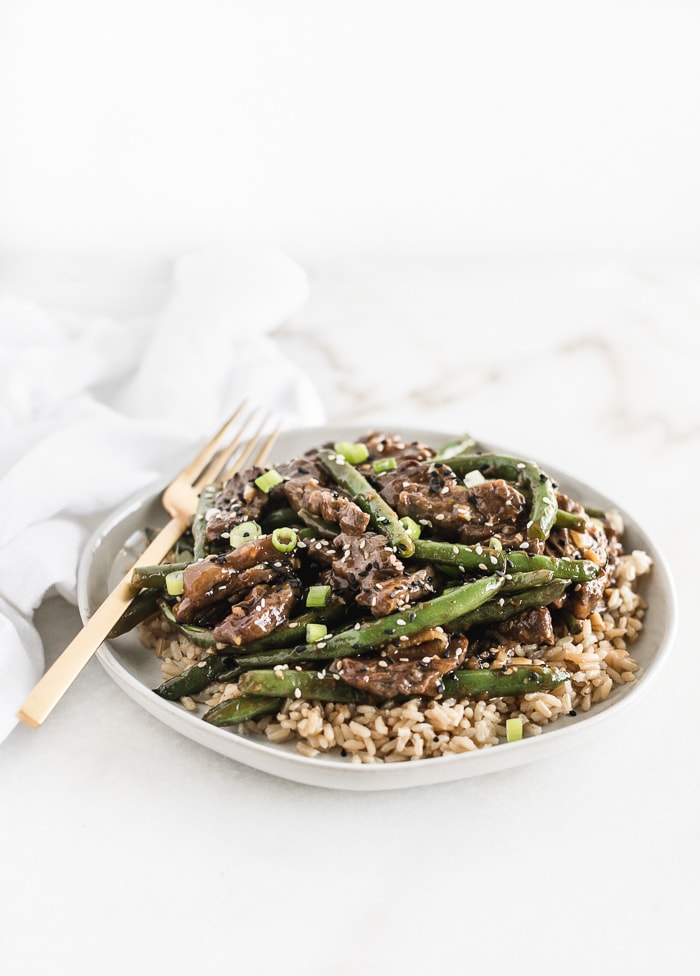 sesame ginger beef and green beans on top of brown rice on a white plate with a gold fork.