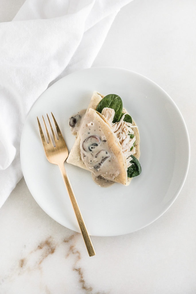 overhead view of a crepe stuffed with chicken topped with mushroom sauce on a white plate with a gold fork.