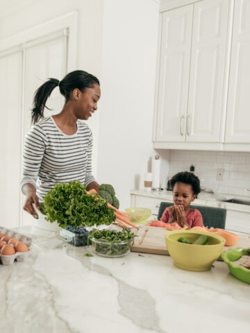 woman and toddler in the kitchen with various food on the counter.