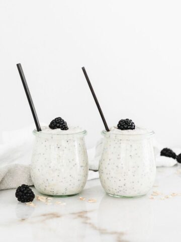 two jars of overnight oats side by side with black spoons in them and blackberries on top.