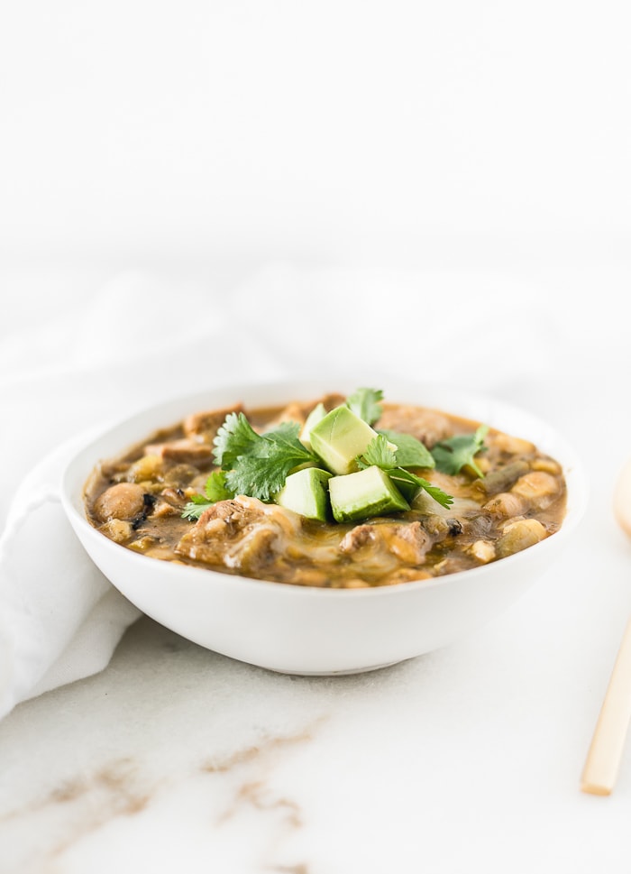 a small white bowl of pork green chile stew topped with cheese and avocado.