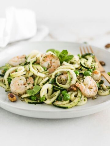zucchini noodles with shrimp, peas and mint pesto on a white plate with a gold fork.