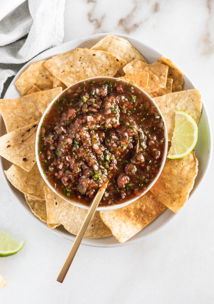 overhead view of a bowl of homemade salsa with a gold spoon in it surrounded by tortilla chips.
