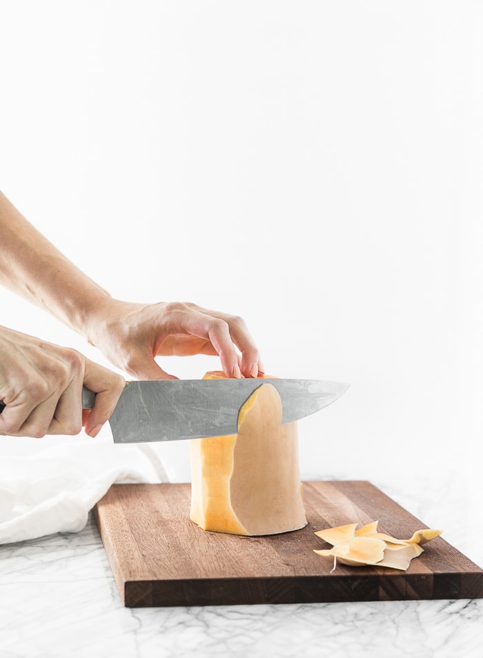 hands using a knife to peel a butternut squash on a wooden cutting board.