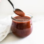 homemade red enchilada sauce in a glass jar with a black spoon dipping some out.