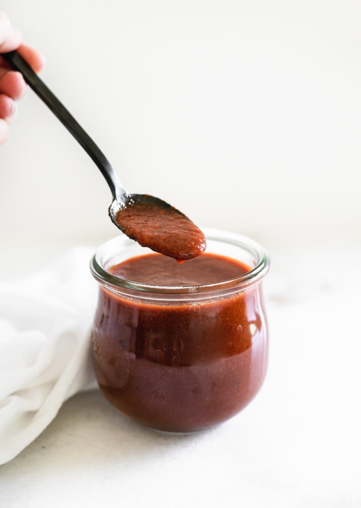 homemade red enchilada sauce in a glass jar with a black spoon dipping some out.