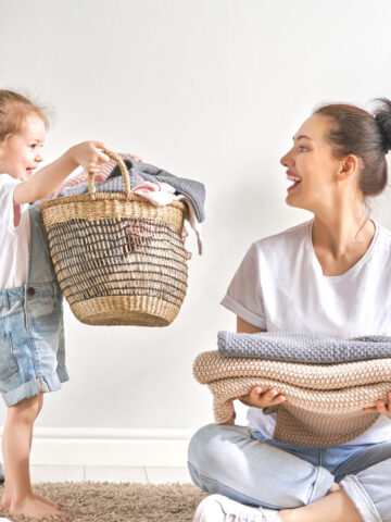 toddler in overalls holding a laundry basket and a woman in a white shirt holding folded blankets.