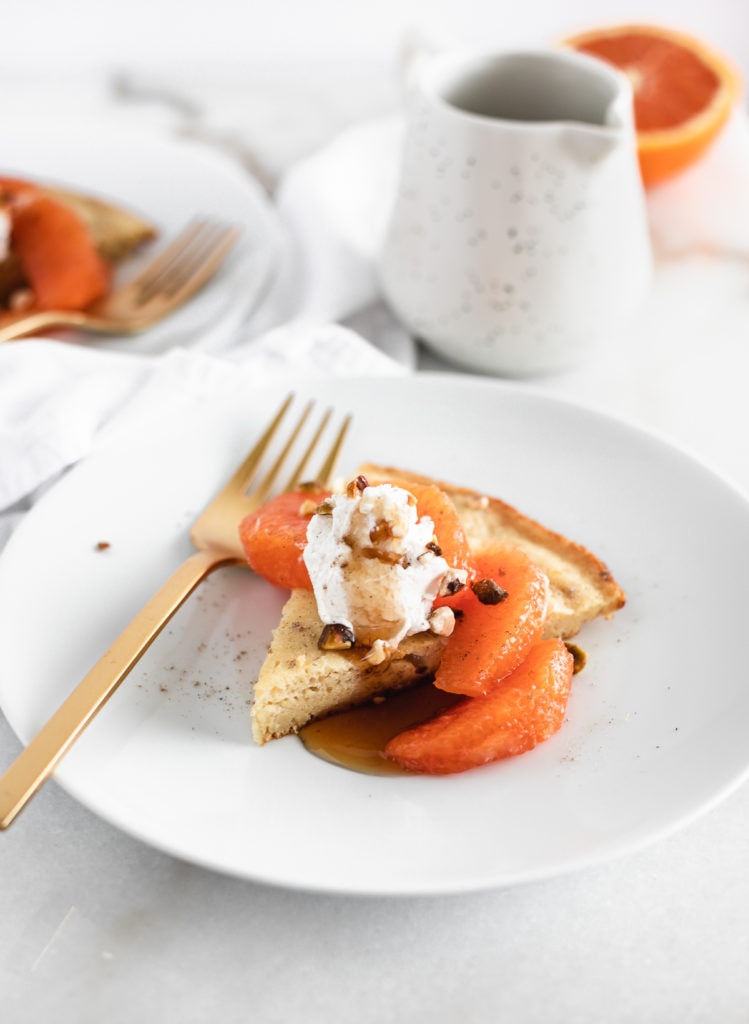 slice of skillet pancake topped with cara cara oranges and whipped cream on a white plate with a gold fork.