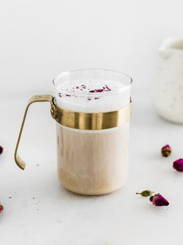 cardamom rose latte in a glass mug with a gold handle surrounded by rose buds.