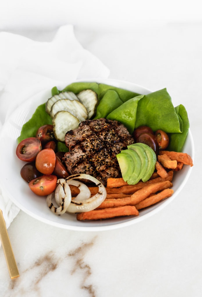 hamburger patty on top of fresh lettuce, tomato, pickles and avocado with sweet potato fries in a bowl. 