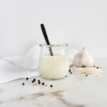 homemade caesar dressing in a glass jar with a black spoon in it, surrounded by garlic and peppercorns.