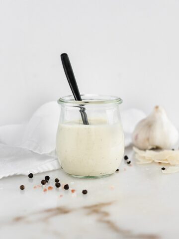 homemade caesar dressing in a glass jar with a black spoon in it, surrounded by garlic and peppercorns.