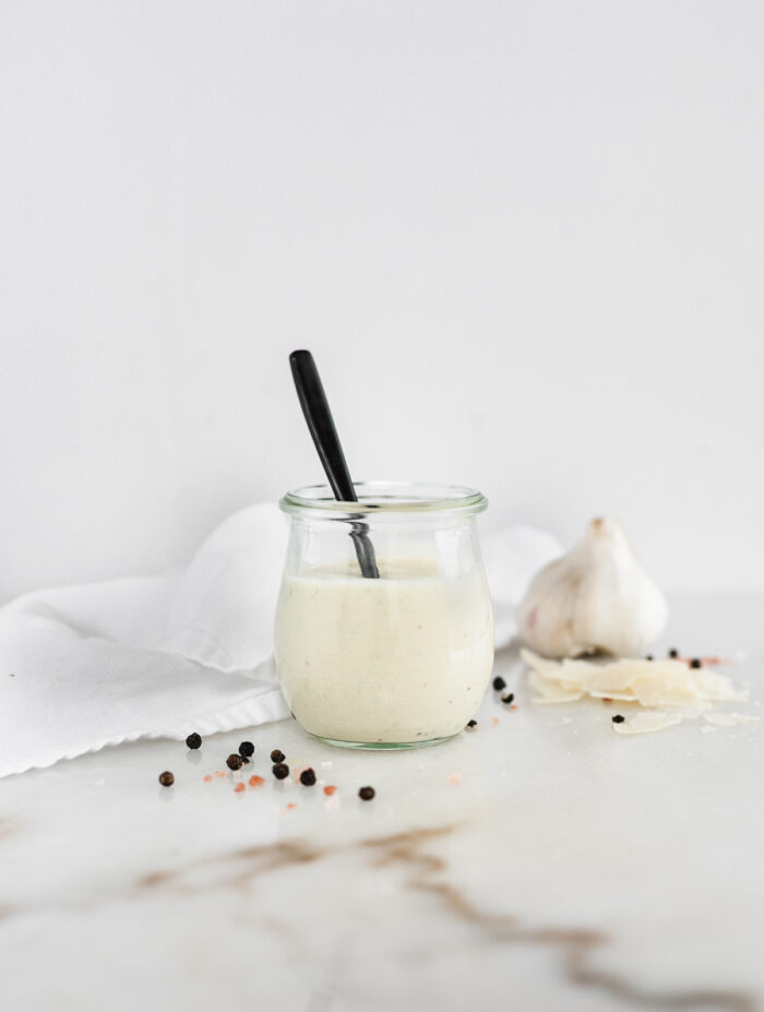 homemade caesar dressing in a glass jar with a black spoon in it, surrounded by garlic and peppercorns.