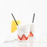 two glasses with coconut orange strawberry chia pudding with black spoons in them and a carafe of orange juice behind them.