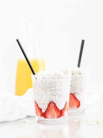 two glasses with coconut orange strawberry chia pudding with black spoons in them and a carafe of orange juice behind them.