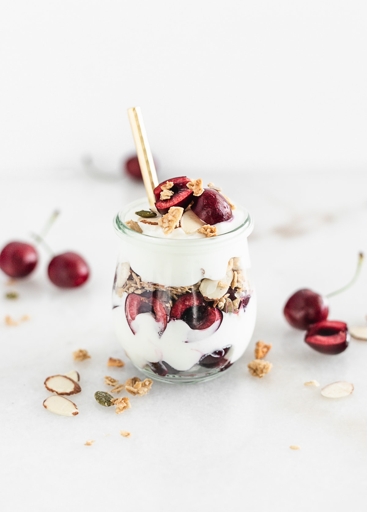 cherry almond yogurt parfait in a glass jar with a gold spoon in it.