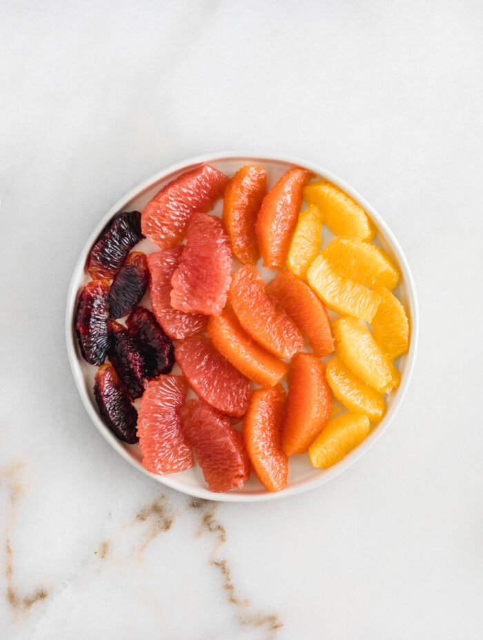 blood orange, grapefruit, cara cara and navel orange segments on a whhite plate against a white marble background.