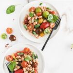 overhead image of two white plates with tomato white bean salad on them with a black fork on one plate and tomatoes and basil around the plates.