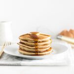 stack of buttermilk pancakes on a white plate with syrup dripping down the front.