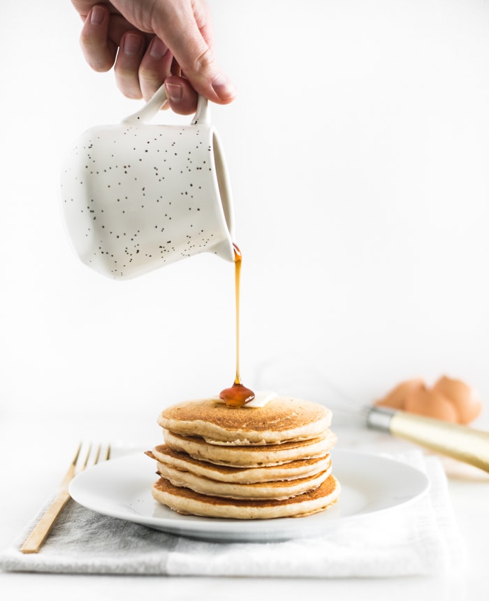 stack of buttermilk pancakes on a white plate with a hand pouring maple syrup on top.