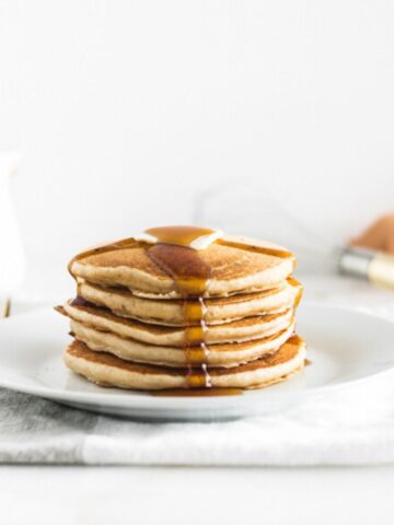 stack of buttermilk pancakes on a white plate with syrup dripping down the front.