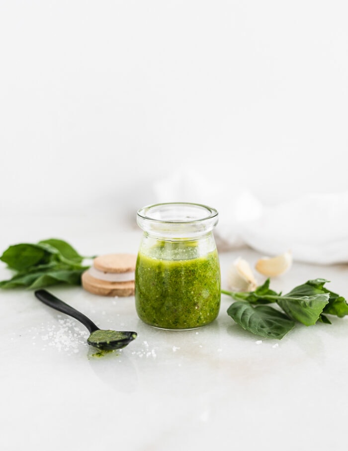 glass jar of basil vinaigrette with a black spoon and basil leaves beside it.