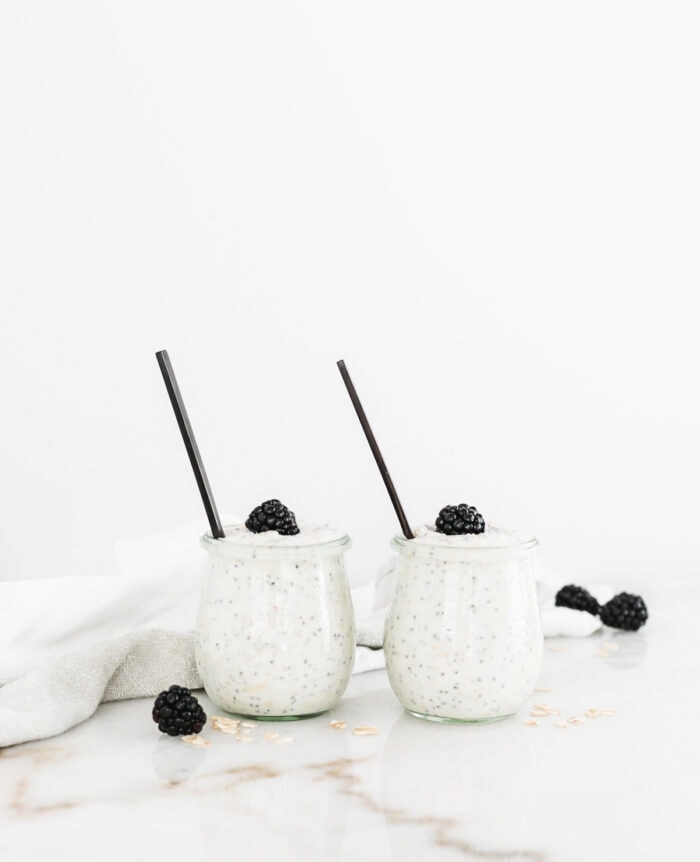 two jars of buttermilk overnight oats sitting side by side with blackberries on top and black spoons in them.