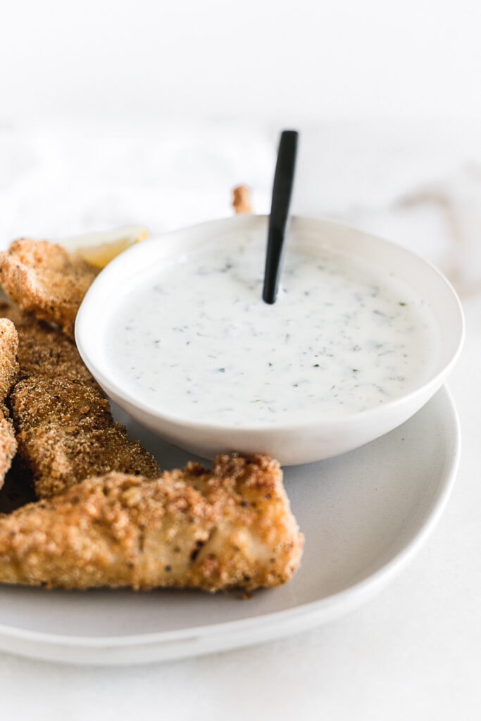 small white bowl of tartar sauce with a black spoon in it on a plate of fried fish.