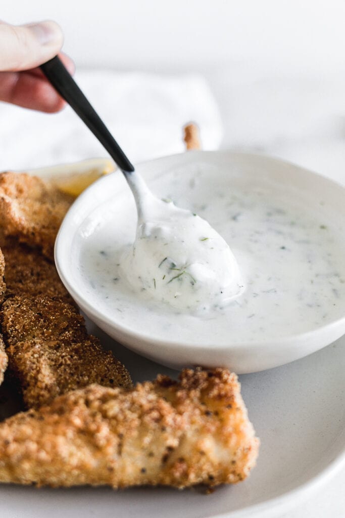 black spoon lifting dill tarter sauce from a small white bowl.