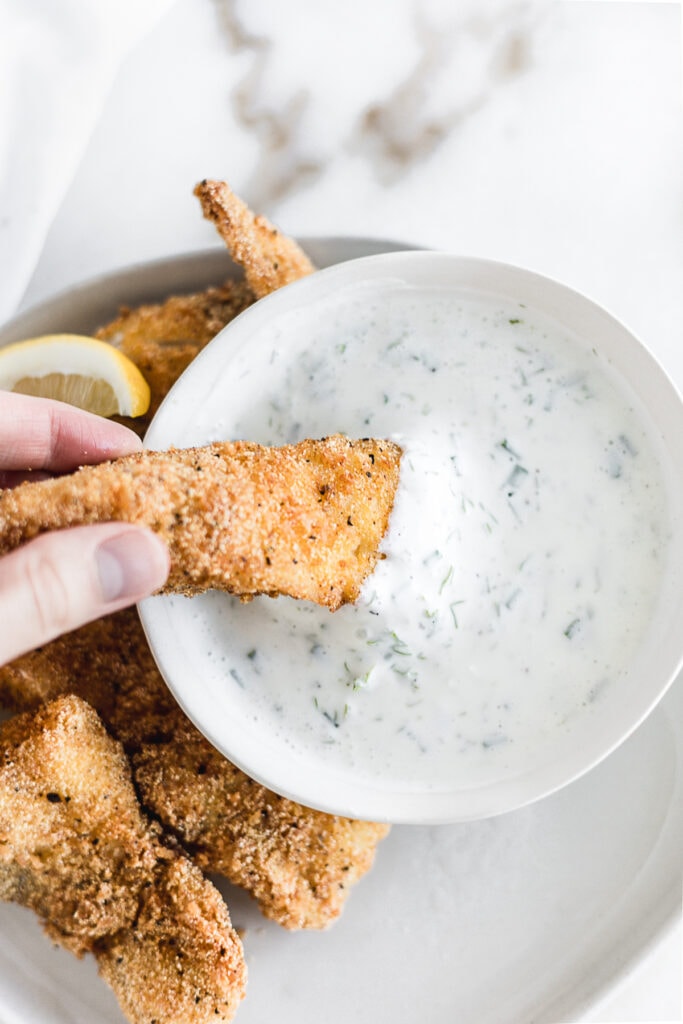 overhead view of a hand dipping a fish filet into a bowl of dill tartar sauce.