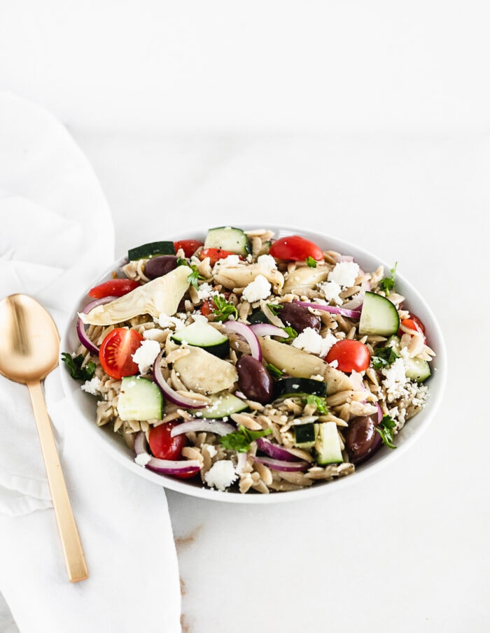 greek orzo pasta salad in a white bowl with a gold spoon beside it.