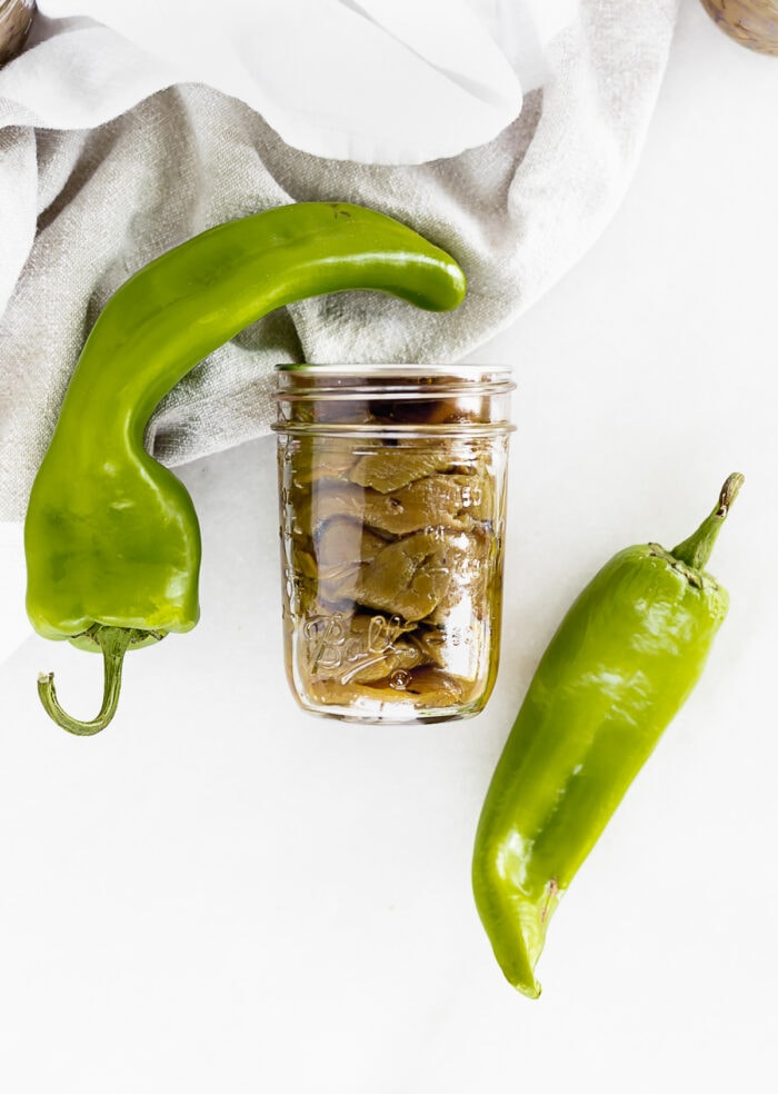 jar of roasted hatch chiles on a white background with two fresh chiles on either side of it.