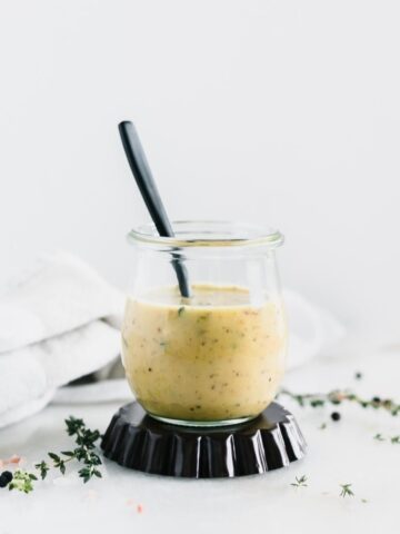 glass jar with maple dijon dressing and a black spoon in it surrounded by thyme and a grey napkin.