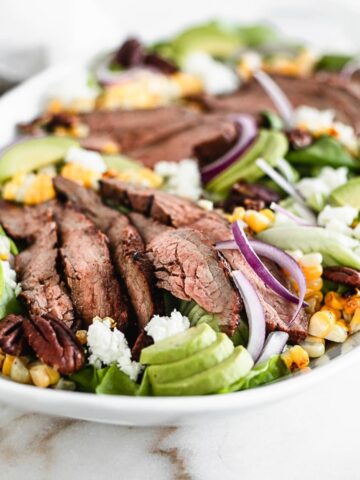 sliced grilled flank steak, avocado, grilled corn and red onion on a salad on a white platter.