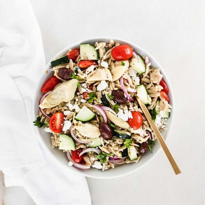 overhead view of greek orzo pasta salad in a white bowl with a gold spoon.