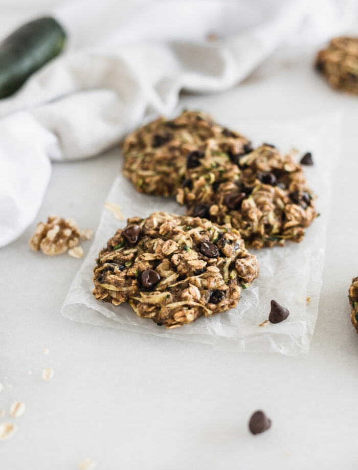 three zucchini oatmeal breakfast cookies on a piece of parchment paper with a white napkin and zucchini in the background.