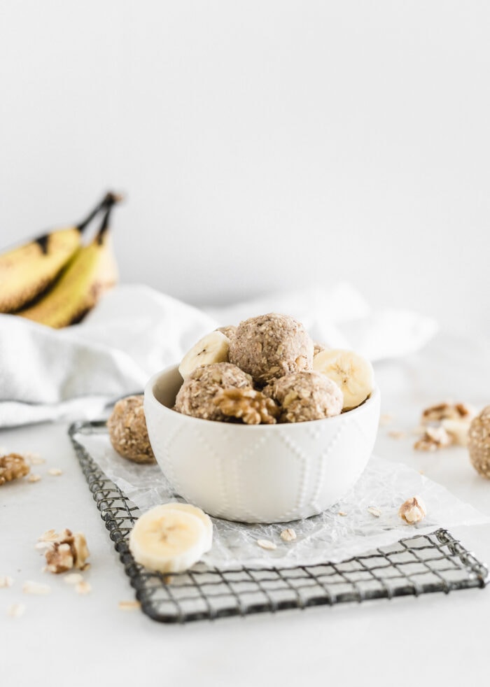 banana bread snack balls in a white bowl on top of a metal rack with banana slices and walnuts around it.