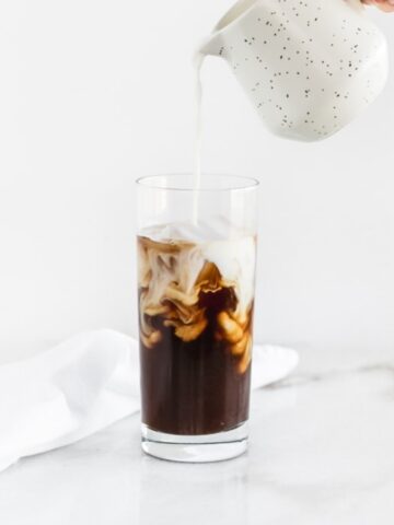 hand pouring milk into a glass of iced coffe.