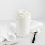 homemade whipped cream in a clear glass with a black spoon beside it.