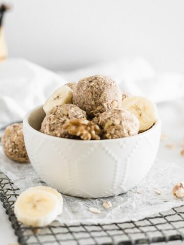 banana bread snack balls in a white bowl on top of a metal rack with banana slices and walnuts around it.
