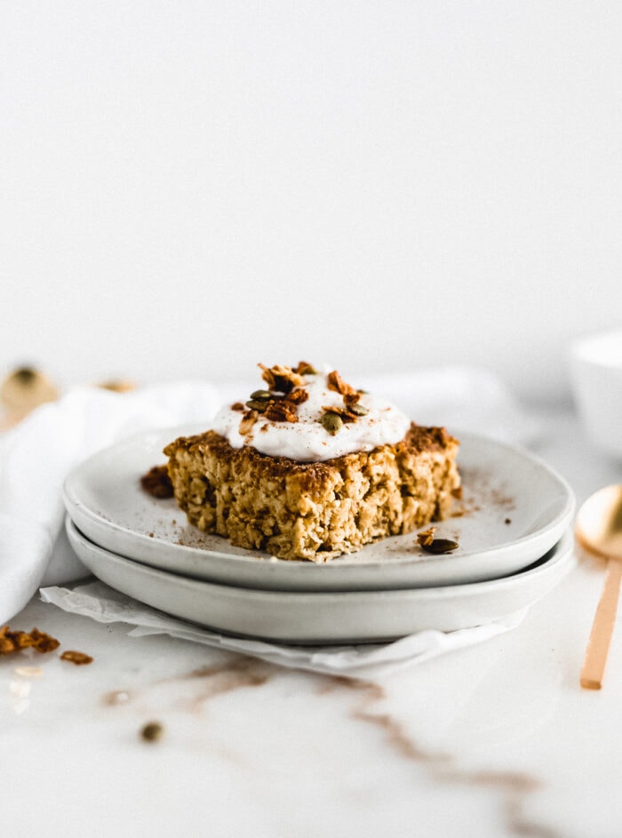 square of pumpkin baked oatmeal topped with yogurt and granola on a stack of grey plates.