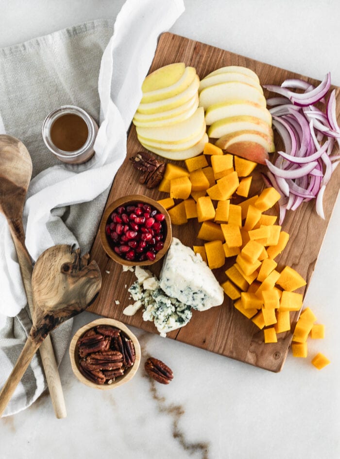 overhead biew of a wooden cuttingboard with cubed pumpkin, sliced apples, sliced red onion, blue cheese, and pomegranate.