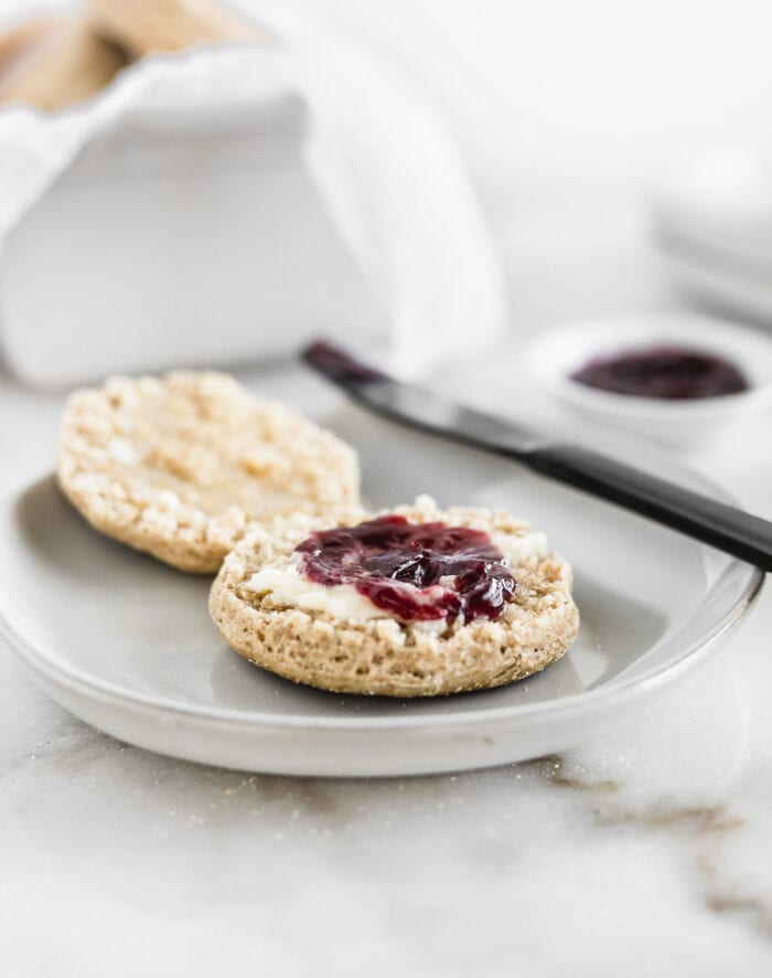 whole wheat sourdough english muffin on a grey plate with butter and grape jam it with a black knife in the background.