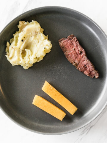 grey plate with a piece of steak, mashed potatoes, and sticks of peaches.