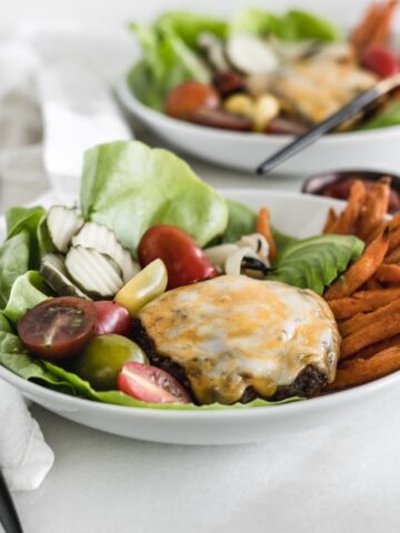 grilled hamburger patty with cheese on top of vegetables and sweet potato fries in a white bowl.